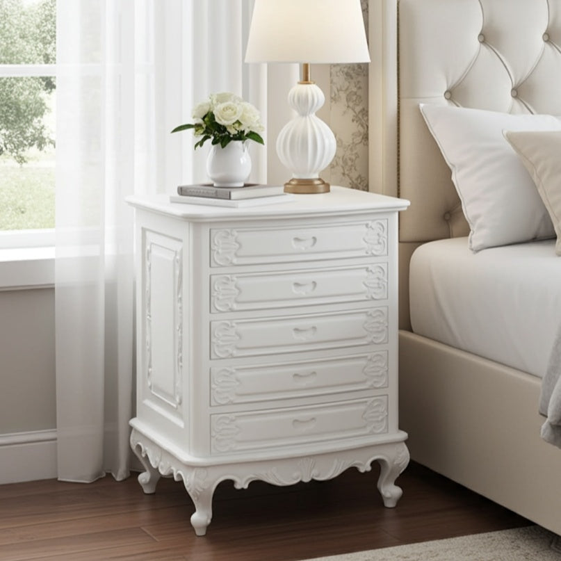 White nightstand with decorative lamp and flowers next to a bed in a bedroom.