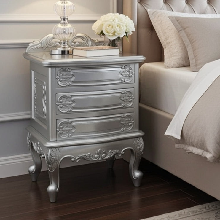 Silver nightstand with decorative elements next to a bed with beige bedding.