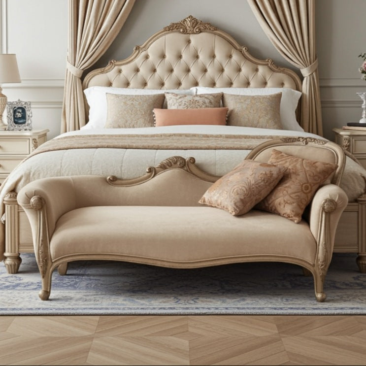 Beige upholstered bed with ornate headboard and chaise lounge in a bedroom setting.